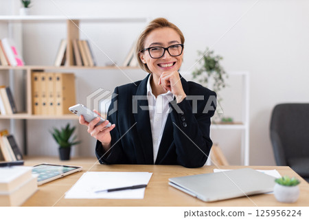 Confident adult woman in formal clothing holding smartphone sitting at desk in office. Workplace features documents, laptop, modern decor. Symbolizes professionalism, communication success in business Confident adult woman in formal clothing holding smartphone sitting at desk in office. Workplace features documents, laptop, modern decor. Symbolizes professionalism, communication success in business 125956224
