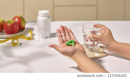 Weight loss pills come in capsule form and have a green shell, helping to improve body shape, placed in the hand, the other hand holds a glass of water to prepare to take the medicine. Weight loss pills come in capsule form and have a green shell, helping to improve body shape, placed in the hand, the other hand holds a glass of water to prepare to take the medicine. 125956262
