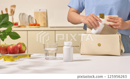 A woman in a blue shirt is taking a pill bottle out of bag. On the dining table are an unlabeled prescription bottle with tight-fitting lid, a glass of water, a plate of apples and a measuring tape. A woman in a blue shirt is taking a pill bottle out of bag. On the dining table are an unlabeled prescription bottle with tight-fitting lid, a glass of water, a plate of apples and a measuring tape. 125956263