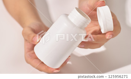 Close-up view of a hand holding unlabeled white open pill bottle, the cap resting in the other hand. The pill box is medium sized, used to hold compressed pills. White background, health supplement. 125956271