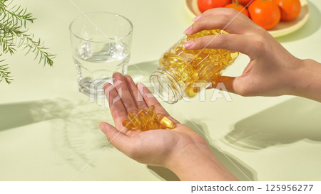 A hand holding an open medicine bottle and pouring out a few pills. A glass of pure water, a plate of tomatoes and a pot plant. The light shone in, casting shadows on the objects on the table. 125956277