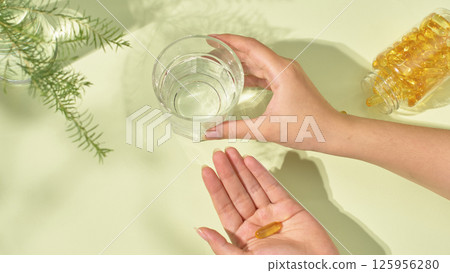A dietary supplement placed in the palm, hand holding a glass of water placed on the table to take medicine. A potted plant on the table with tiny leaves. Shadows of objects cast on the table. 125956280