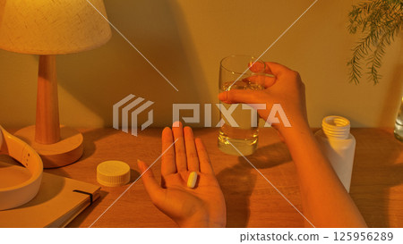 The patient takes medication before going to bed with vitamin capsules placed in the palm of one hand the other hand holding a glass of water ready to drink. The daily dose is placed on the table. The patient takes medication before going to bed with vitamin capsules placed in the palm of one hand the other hand holding a glass of water ready to drink. The daily dose is placed on the table. 125956289