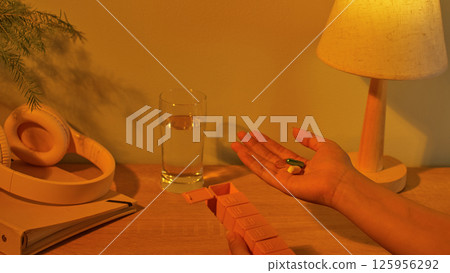 A daily pillbox with the first compartment open. The person in the photo holds two pills, ready to take before bed. Headphones are placed on a diary. The lighting in the room is a soft yellow. 125956292