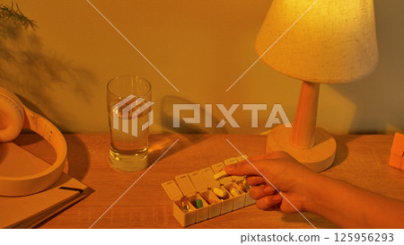 Capsules of different colors are arranged in compartments corresponding to each day. Light is emitted from a nightlight in the room, and on the table are headphones, a glass of water, and a notebook. 125956293