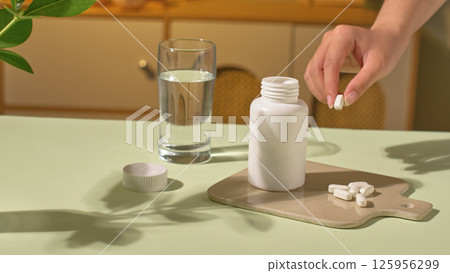 A hand is picking up a pill, some capsules placed on the tray, next to it is an open medicine bottle, displayed on a light brown tray. A glass of pure white water and a pot of green plants. A hand is picking up a pill, some capsules placed on the tray, next to it is an open medicine bottle, displayed on a light brown tray. A glass of pure white water and a pot of green plants. 125956299