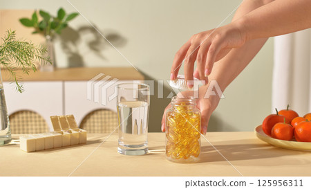A medicine bottle and glass of water are placed on the table. One hand holds the medicine bottle, the other hand holds the open cap. Medicine box placed on the table with a few compartments open. A medicine bottle and glass of water are placed on the table. One hand holds the medicine bottle, the other hand holds the open cap. Medicine box placed on the table with a few compartments open. 125956311