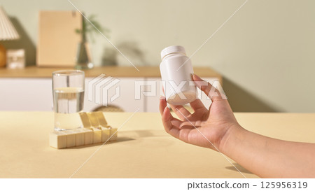 The hand holding the medicine bottle is made of synthetic plastic, safe for health, to preserve the capsules during use. Behind are glass and beige medicine boxes with compartments divided by day. The hand holding the medicine bottle is made of synthetic plastic, safe for health, to preserve the capsules during use. Behind are glass and beige medicine boxes with compartments divided by day. 125956319