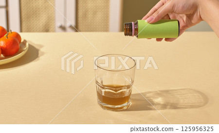 The action of pouring collagen from a bottle into a glass cup placed on the table. Collagen water is brown, liquid, contains many vitamins and nutrients that are good for health. Healthcare concept. 125956325