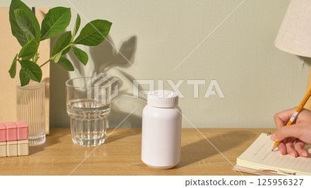 Mockup of a white drug bottle, used for advertising medical products. On the desk there is a glass of water, a vase, a branch and a notebook. The student is using a pencil to write on the notebook. Mockup of a white drug bottle, used for advertising medical products. On the desk there is a glass of water, a vase, a branch and a notebook. The student is using a pencil to write on the notebook. 125956327
