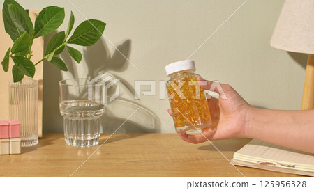 A drug bottle containing capsules full of nutrients is held in the air by someone's hand. On the table was a glass of water. A branch was placed in the bottle. White background with leaves shadows. A drug bottle containing capsules full of nutrients is held in the air by someone's hand. On the table was a glass of water. A branch was placed in the bottle. White background with leaves shadows. 125956328