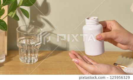 Hand is holding an opened medicine bottle, pouring the pills into the other hand. Water helps the patient take the medicine more easily. The white wall with the shadow of the leaves on the wall. 125956329
