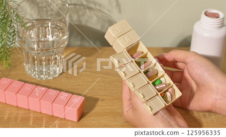 Two hand holding a beige personal daily pill box, inside each compartment are colorful pills, arranged according to the doctor's prescription. Another pink pill box is placed on the table. 125956335