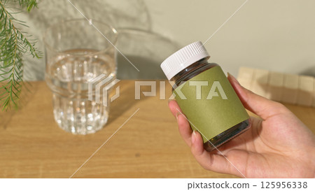 A hand is holding a bottle of dietary supplements with a light blue outer shell and a white cap, raised towards the camera. A glass of water is placed on a light brown wooden table. White background A hand is holding a bottle of dietary supplements with a light blue outer shell and a white cap, raised towards the camera. A glass of water is placed on a light brown wooden table. White background 125956338