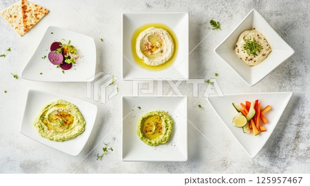 Vibrant hummus varieties spreading across white plates, accompanied by raw vegetable slices, presenting colorful mediterranean inspired healthy eating arrangement 125957467