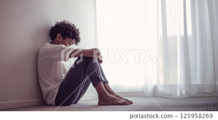 A teenage Black boy with short curly hair sits on the floor of his bedroom, gazing into space. Natural light gently illuminates his face while the untidy room fades into the background 125958269