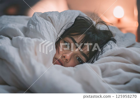 Teenage Latina girl lies in bed, her weary eyes peering from under a thick blanket. The soft evening light reveals a dim room, capturing a moment of introspection and emotional fatigue 125958270
