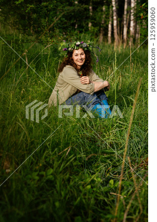 Woman in casual clothes and flower crown sitting in the grass, smiling and holding berry. Rural eco-lifestyle, summer joy and unplugged simplicity Woman in casual clothes and flower crown sitting in the grass, smiling and holding berry. Rural eco-lifestyle, summer joy and unplugged simplicity 125959060