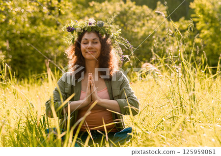 Woman meditating in the wild grass under sunlight, wearing floral wreath and casual clothing. Scene of unplugged mindfulness, eco-energy and inner balance 125959061