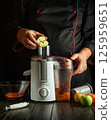 A skilled chef is preparing a fresh juice by placing slices of apple into a juicer. The warm kitchen is filled with fruits, creating a cozy atmosphere during the evening 125959651