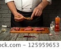 A chef prepares raw meat by carefully sprinkling seasoning from a spoon, showcasing culinary skills. The kitchen is well-lit, enhancing the focus on the fresh ingredients 125959653