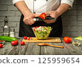 A chef is slicing tomatoes while preparing a fresh salad in a contemporary kitchen. Colorful vegetables like cucumbers and radishes surround the chef, creating a vibrant atmosphere 125959654