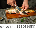 A chef skillfully fillets a fresh fish on a wooden cutting board in a cozy kitchen. The hands show attention to detail using a sharp knife. Fresh herbs and spices add color nearby 125959655