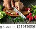 A person is finely slicing radishes using a sharp knife on a wooden cutting board. Surrounding the board are fresh greens and herbs, showcasing a vibrant and healthy preparation 125959656