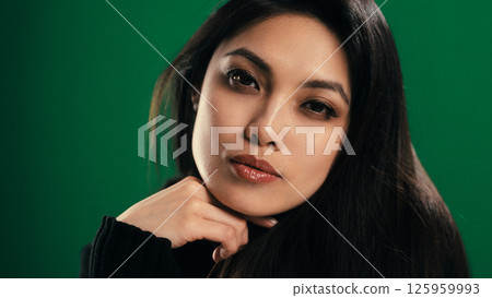 Portrait of Confident Young Asian Woman with Hand on Chin Against Green Backdrop 125959993