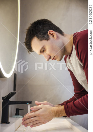 Young man thoroughly washes his hands under running water, focusing on proper hygiene and cleanliness in a modern bathroom. Young man thoroughly washes his hands under running water, focusing on proper hygiene and cleanliness in a modern bathroom. 125962023