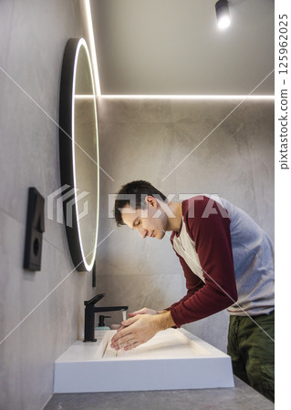 Young man calmly washes his hands at the sink, maintaining good hygiene and healthy habits in a bright, modern bathroom. 125962025