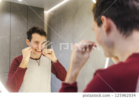 Young man stands at the bathroom mirror flossing his teeth, smiling while practicing effective oral hygiene as part of a healthy daily routine. Young man stands at the bathroom mirror flossing his teeth, smiling while practicing effective oral hygiene as part of a healthy daily routine. 125962034