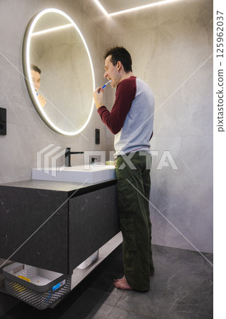 Young man stands barefoot in front of a bathroom mirror, brushing his teeth as part of a healthy hygiene routine in a modern and clean space. Young man stands barefoot in front of a bathroom mirror, brushing his teeth as part of a healthy hygiene routine in a modern and clean space. 125962037