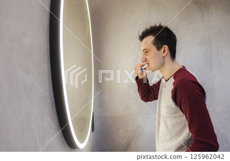 Young man in a casual sweatshirt brushing his teeth in front of a round illuminated mirror in a modern bathroom, smiling slightly as he looks at his reflection. Young man in a casual sweatshirt brushing his teeth in front of a round illuminated mirror in a modern bathroom, smiling slightly as he looks at his reflection. 125962042