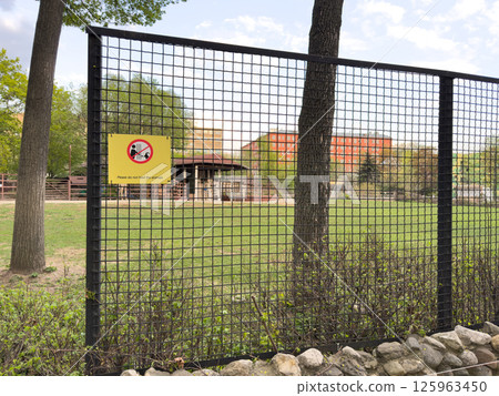Wire fence with yellow warning sign in zoo area instructing visitors not to feed animals, surrounded by green grass, trees and enclosures, serving as protective barrier and rule enforcement element Wire fence with yellow warning sign in zoo area instructing visitors not to feed animals, surrounded by green grass, trees and enclosures, serving as protective barrier and rule enforcement element 125963450