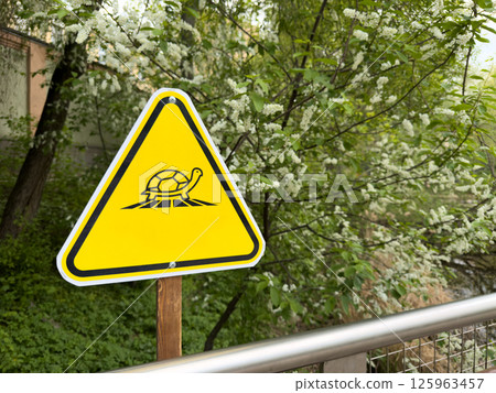 Bright yellow triangular warning sign with turtle icon stands in green park zone, serving as clear visual symbol in public space, combining environmental education with modern graphic design Bright yellow triangular warning sign with turtle icon stands in green park zone, serving as clear visual symbol in public space, combining environmental education with modern graphic design 125963457