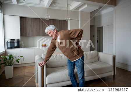 Grimacing old man in pain suffers sudden back spasm due to spinal hernia, standing alone by sofa 125968357