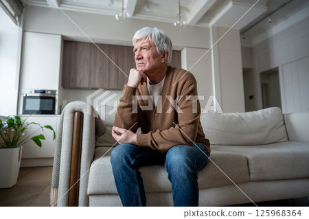 Thoughtful struggling elderly man rests fist against cheek, looking out window alone on sofa Thoughtful struggling elderly man rests fist against cheek, looking out window alone on sofa 125968364