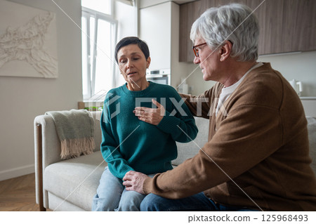Wife health issues. Mature woman grab chest struggles for air comforted on couch by supportive man 125968493