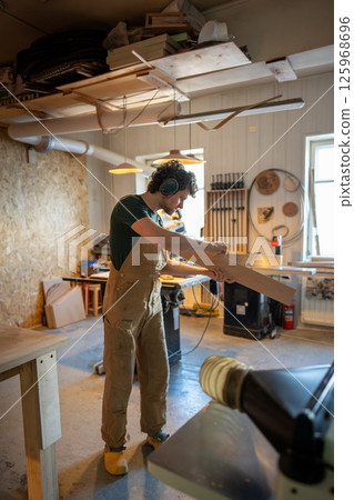 Focused handyman with earmuffs measuring wood board thickness with calipers in furniture workshop 125968696