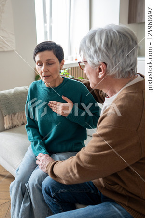 Asthma breathing, heart attack, Struggling mature woman clutches chest while man supports on sofa Asthma breathing, heart attack, Struggling mature woman clutches chest while man supports on sofa 125968697