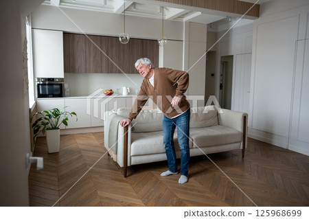 Elderly man backache. Senior male barely standing from sofa amid kidney disease and nerve damage Elderly man backache. Senior male barely standing from sofa amid kidney disease and nerve damage 125968699