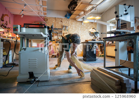 Focused busy carpenter woodworker clean floor with industrial vacuum hose after using planer machine 125968960