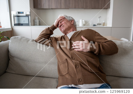 Elderly pensioner lies on couch with chest pain, tearful expression suggesting cardiac emergency 125969120