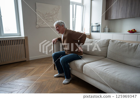 Lonely unhealthy elderly man holds aching back unable to rise from couch due to spinal problems 125969347