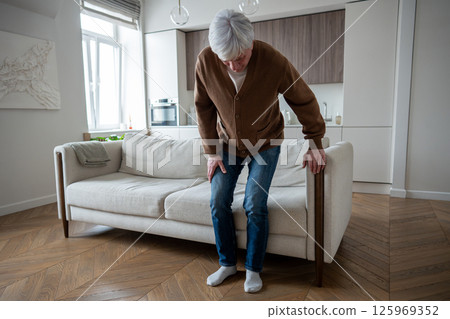 Elderly man with joint dysfunction painfully rises from couch clutching arthritic knee at home  125969352