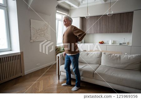 Sad elderly man painfully straightens hunched back after rising from couch with severe spinal pain Sad elderly man painfully straightens hunched back after rising from couch with severe spinal pain 125969356