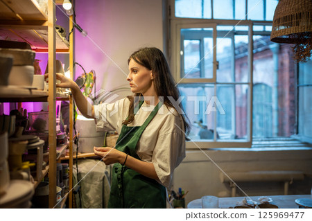 Woman places completed ceramic items on shelves in workshop of gift store, preparing for drying Woman places completed ceramic items on shelves in workshop of gift store, preparing for drying 125969477