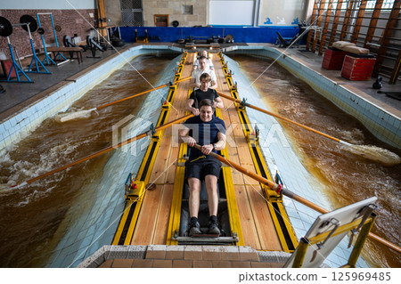 Concentrated tired athletes stroking on rowing simulator. Synchronization, water aquatic sports Concentrated tired athletes stroking on rowing simulator. Synchronization, water aquatic sports 125969485