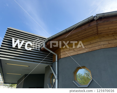 Modern public restroom building with large WC letters on metal facade and wooden roof detail under blue sky, combining practical function with clean architectural design in outdoor urban space 125970016